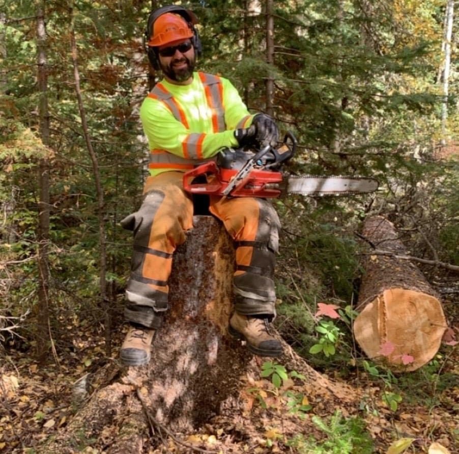default-18 A bearded man with safety gear poses with a chainsaw on a stump, next to a fallen tree.
