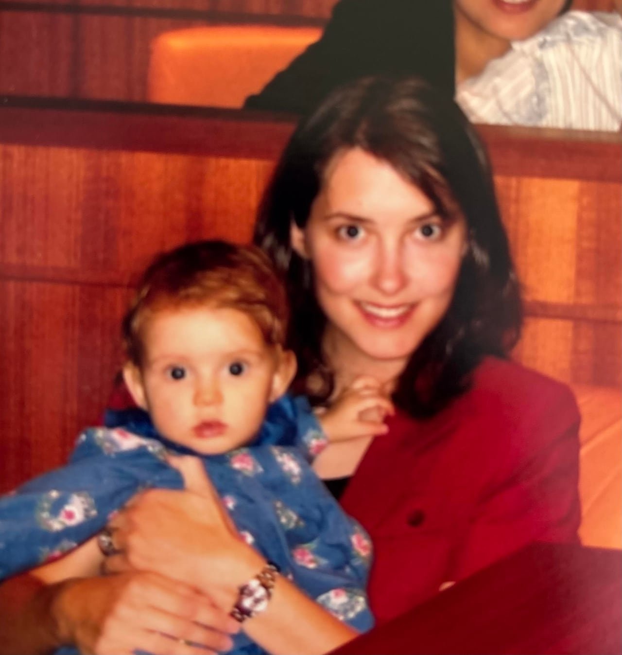 A smiling woman in a red suit holds a baby girl in her arms.