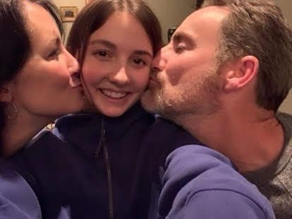 A teenage girl snaps a selfie as she is kissed on her cheeks by a woman on the left and a man on the right.