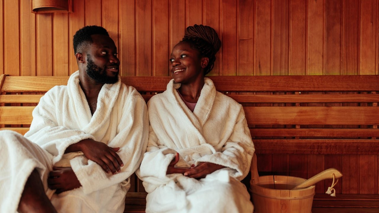 default-547 A couple wearing bath robes, sitting on a bench in a wood-panelled sauna, smiling at one another.
