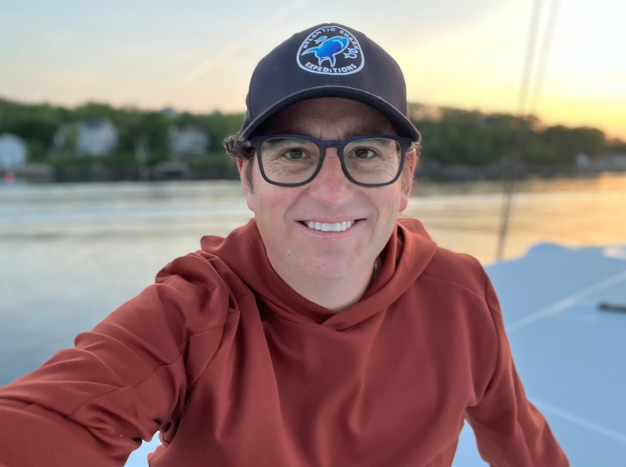 default-60 A smiling man with square glasses. He is wearing a baseball hat and an orange hoodie sitting on a boat.