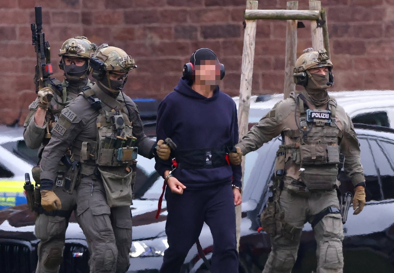 default-98 An individual wearing headphones, a ballcap, and a hoodie, with blurred facial features, being escorted by three police officers in camouflage attire and carrying firearms.